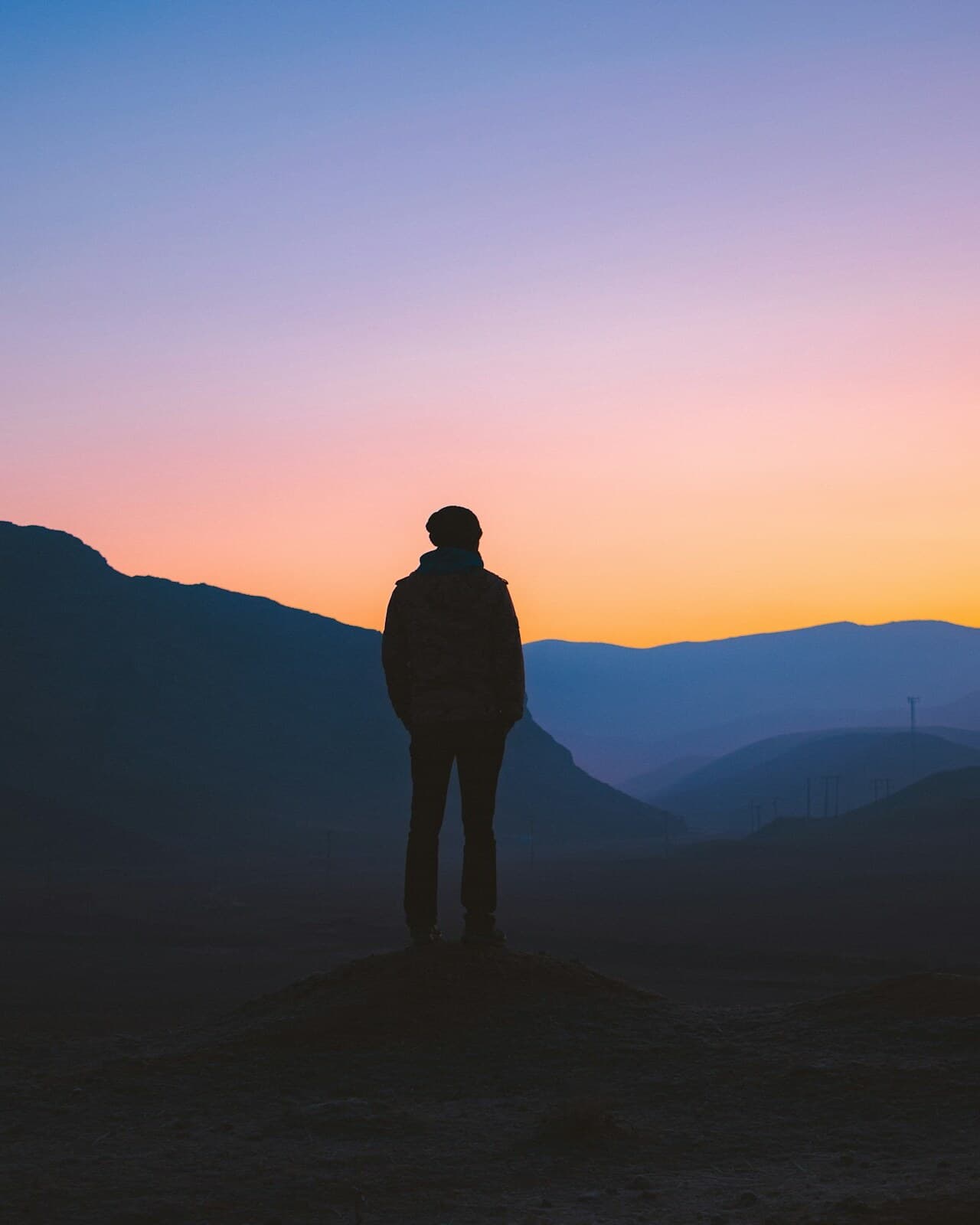 A figure standing at dawn looking out over layered mountains and a gradient sky