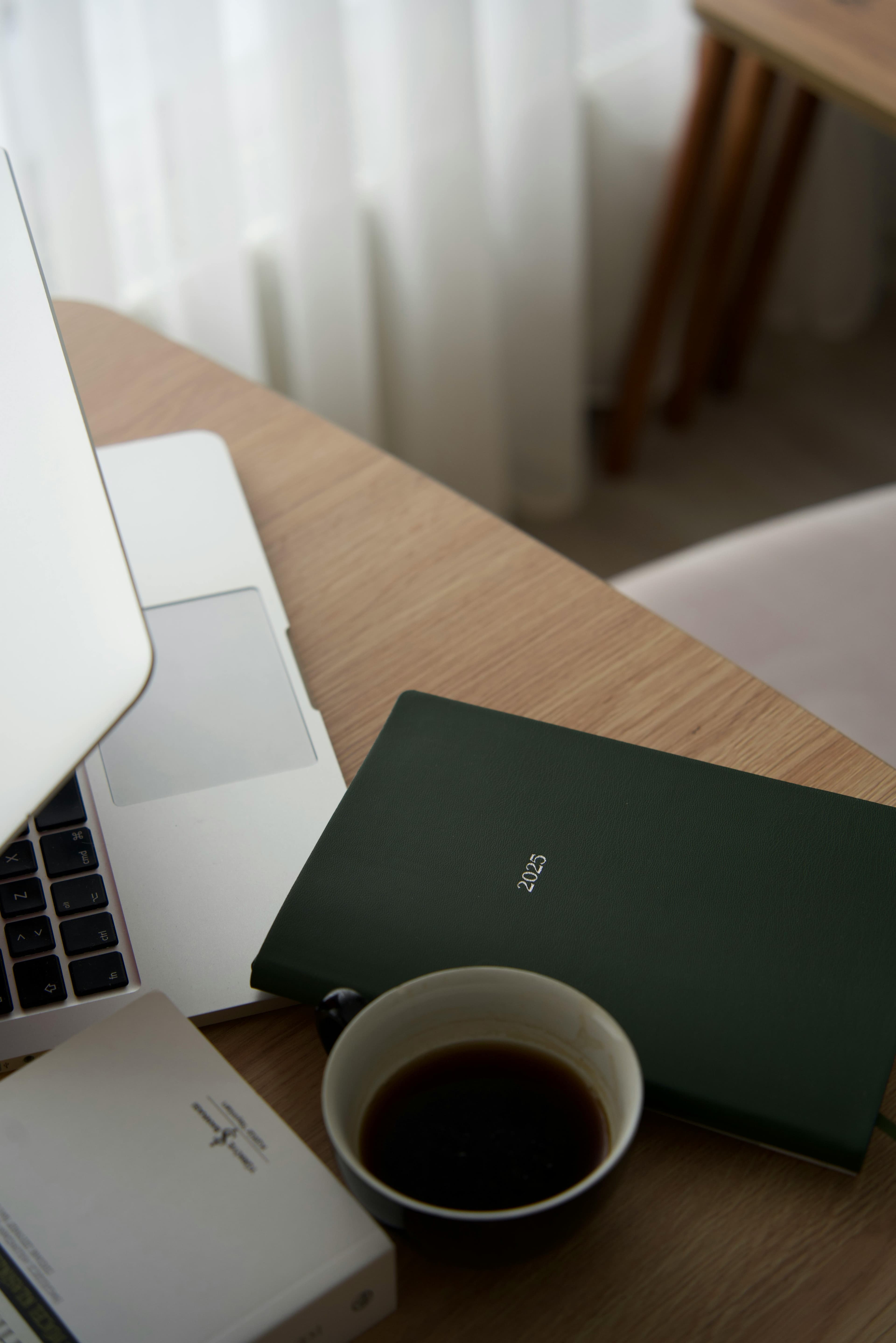 A laptop on a wooden desk beside a green notebook and a coffee cup in soft window light